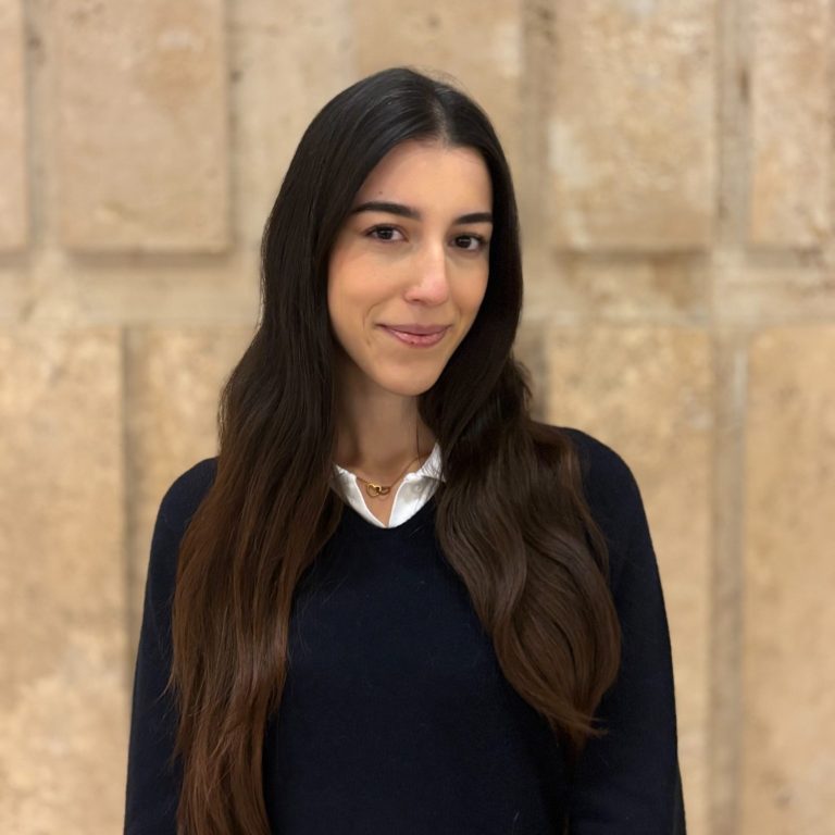 (c) Ester Shahini Junge Frau mit langen, glatten Haaren, trägt einen dunkelblauen Pullover und lächelt.