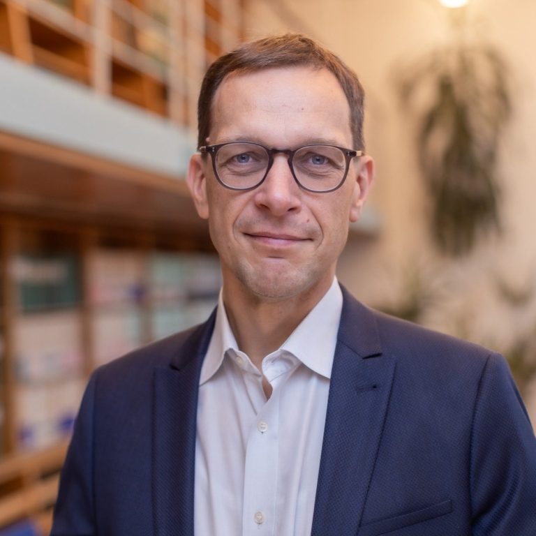 (c) Nils Eisfeld Mann mit Brille in Anzug, freundlicher Blick, Hintergrund mit Regalen.