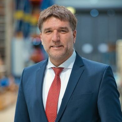 Mann in Anzug mit Krawatte, lächelt vor einer industriellen Hintergrundszene.