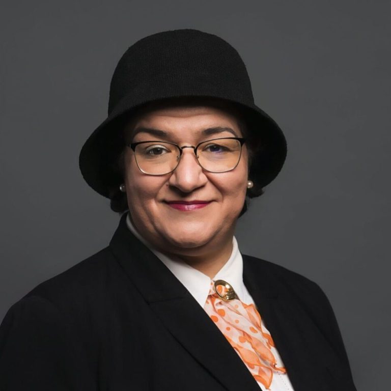 (c) MST / TUC Eine lächelnde Person trägt einen schwarzen Hut und Anzug mit einem orangefarbenen Schal.